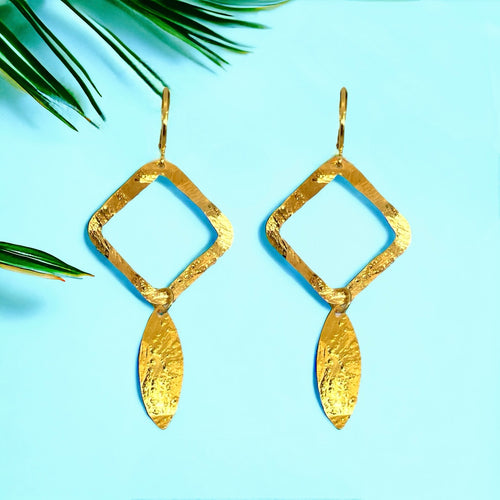 Boucles d'oreilles dorées avec pendentif en forme de losanges et pointes, sur fond bleu et feuilles de palmier