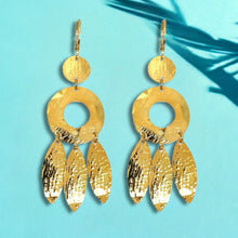 Charger l&#39;image dans la galerie, Longues boucles d&#39;oreilles dorées à l&#39;or fin en métal martelé, de style ethnique bohème chic, avec leurs pampilles en forme de plumes, sur fond bleu et feuilles de palmier
