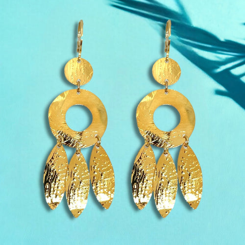 Longues boucles d'oreilles dorées à l'or fin en métal martelé, de style ethnique bohème chic, avec leurs pampilles en forme de plumes, sur fond bleu et feuilles de palmier