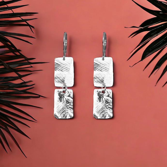 Boucles d'oreilles pendantes composées de 2 rectangles en métal martelé argentées, sur fond orangé avec des feuilles de palmier