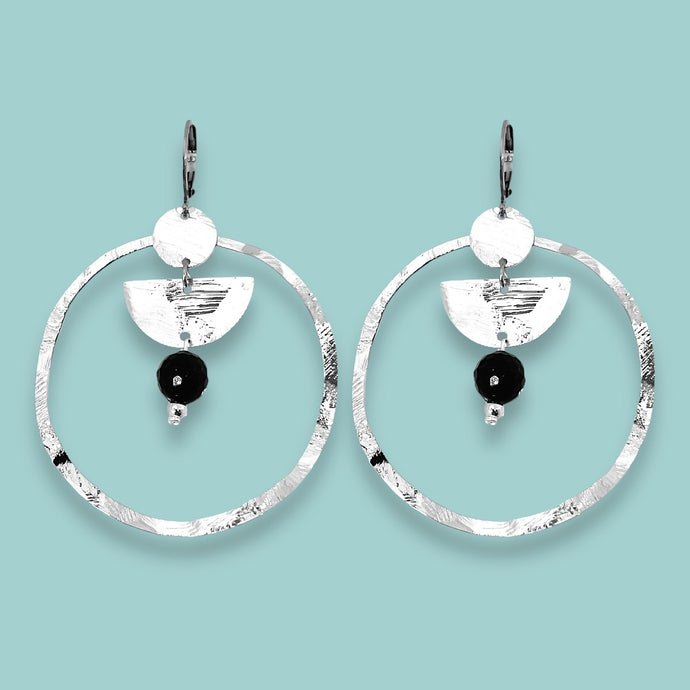 Grandes boucles d'oreilles créoles en métal martelé argenté et pierres naturelles de tourmaline noire accrochée à une demi-lune, sur fond vert pastel