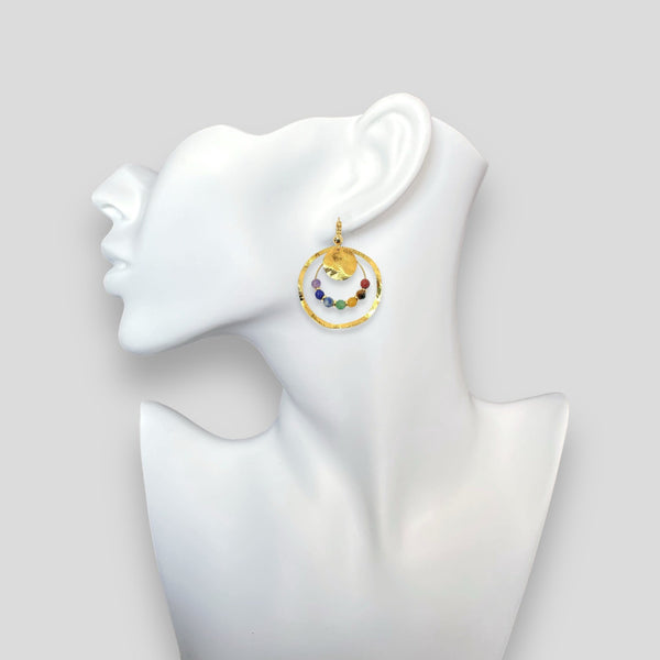 Grandes boucles d'oreilles créoles dorées pendantes longues rondes avec double anneau et petites pierres naturelles multicolores des 7 Chakras, portées à l'oreille sur un buste blanc