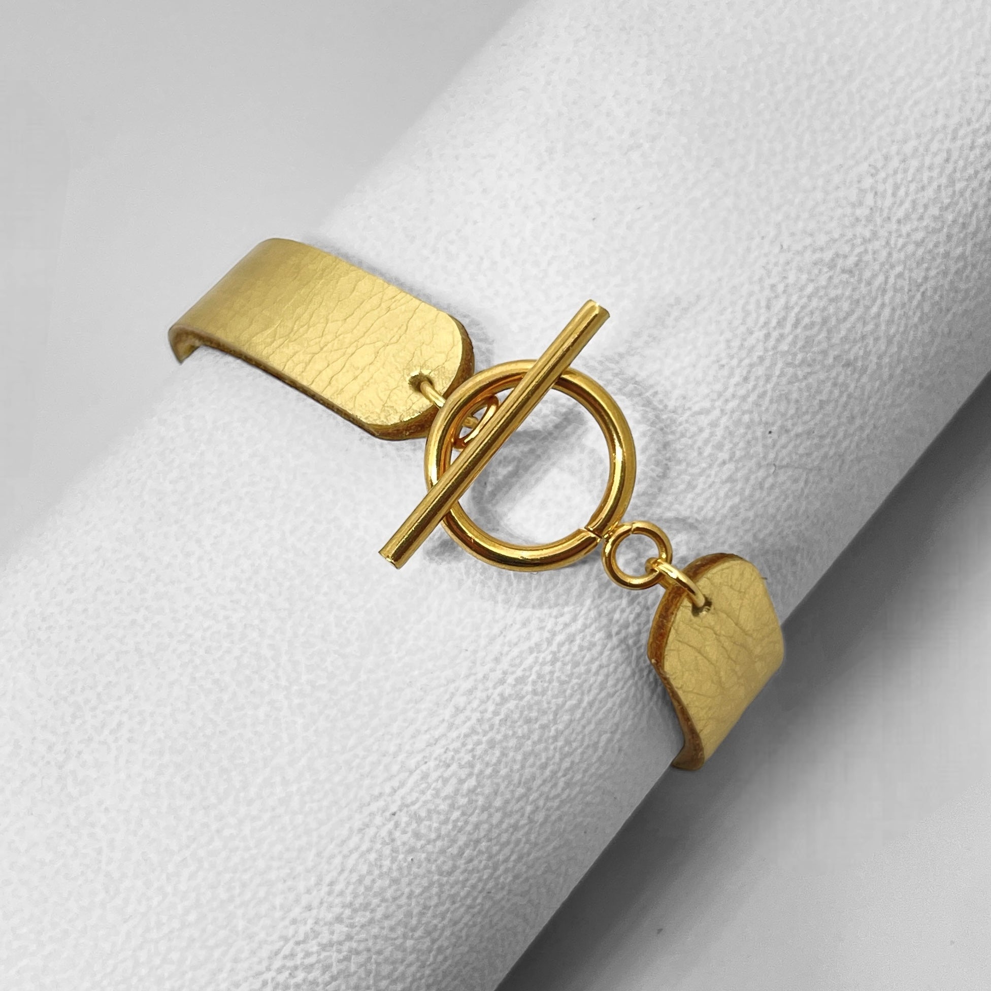 Bracelet en cuir doré brillant vu de dessous avec fermoir en T doré, enroulé autour d'un rouleau de cuir blanc