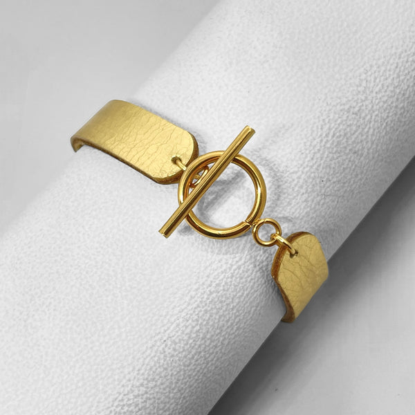 Bracelet en cuir doré brillant vu de dessous avec fermoir en T doré, enroulé autour d'un rouleau de cuir blanc