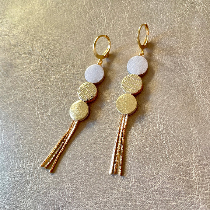 Longues et fines boucles d'oreilles Nune à franges dorées avec 3 petits disques de cuir recyclé blanc cassé nacré, doré façon serpent et doré, sur fond beige doré. Modèle Janis