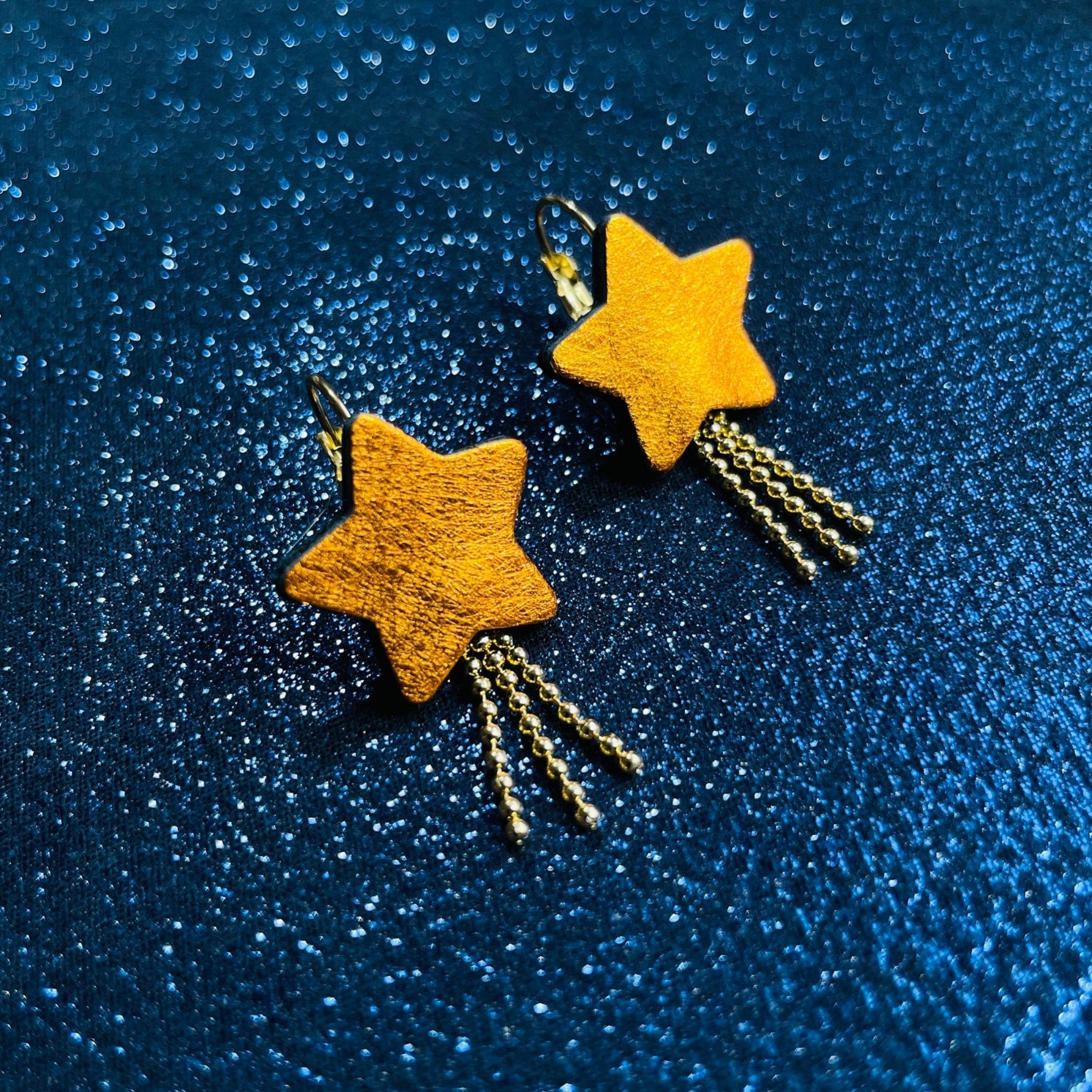 boucles d'oreilles dorées Nune modèle Stella en forme d'étoiles, en cuir orange métallisé, sur fond noir paillettes