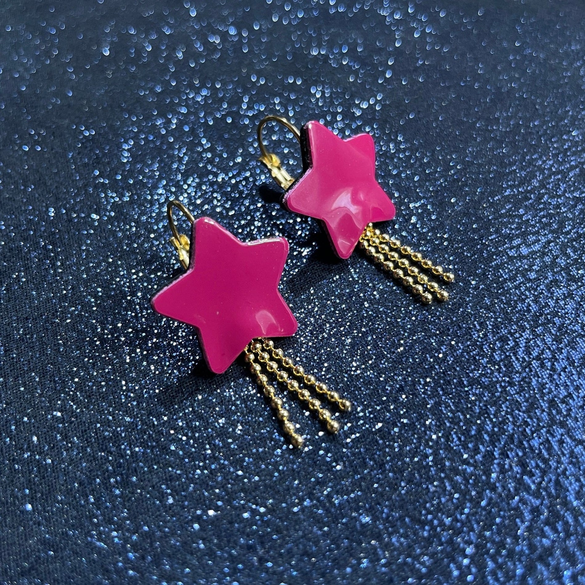 boucles d'oreilles dorées Nune modèle Stella en forme d'étoiles, en cuir rose fushia vernis, sur fond noir paillettes
