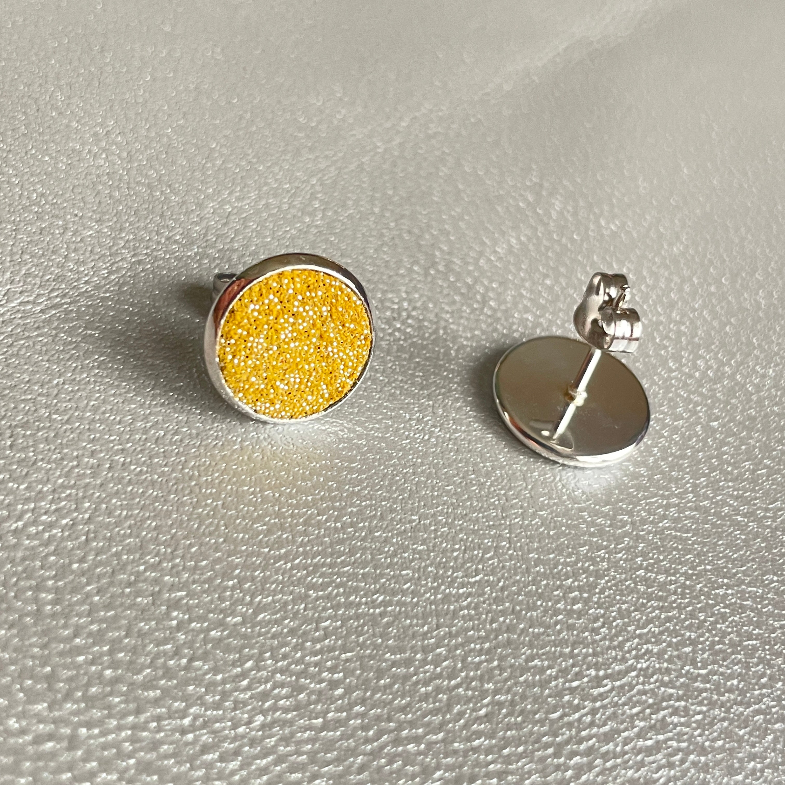 Puces d'oreilles argentées rondes Nune modèle Ilis argenté pailleté avec pastilles de cuir safran pailleté argent, sur fond de cuir gris métallisé