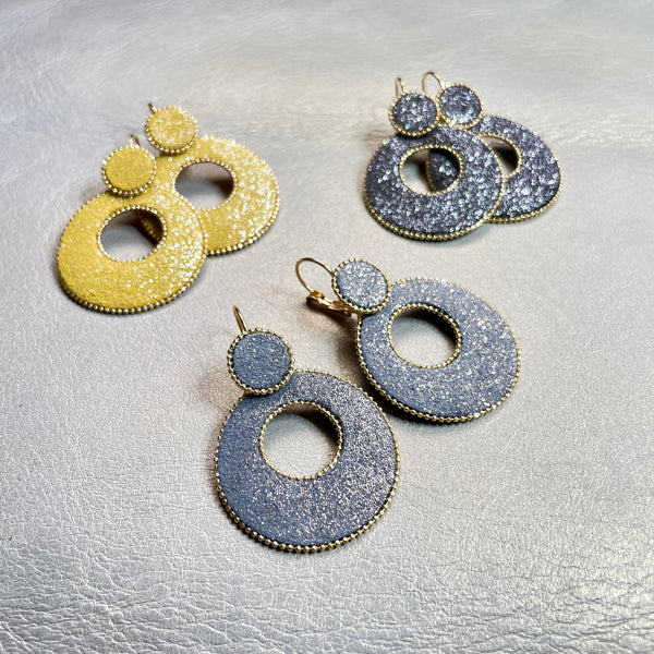 boucles d'oreilles créoles Nune modèle Céleste en forme de cercles en cuir gris pailleté or, safran pailleté argent et anthracite pailleté argent cerclées de chaîne dorée, sur fond beige doré
