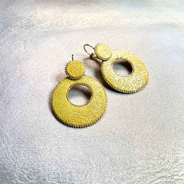 boucles d'oreilles créoles Nune modèle Céleste en forme de cercles en cuir safran pailleté argent, cerclées de chaîne dorée, sur fond beige doré