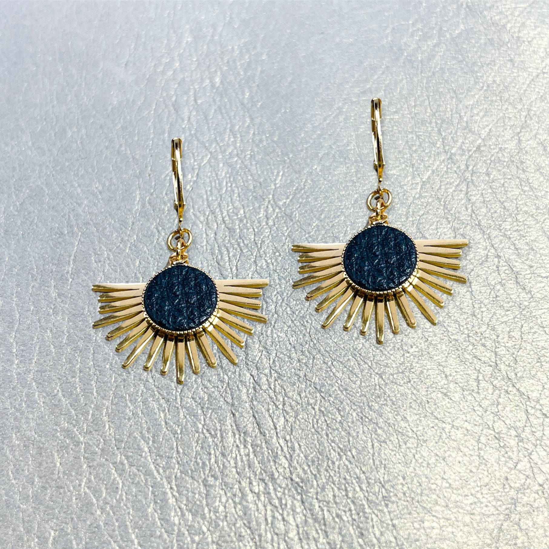 Petites boucles d'oreilles dorées Nune modèle Miria en acier inoxydable et laiton doré à l'or fin avec serti en cuir noir grainé, posées sur du cuir beige doré