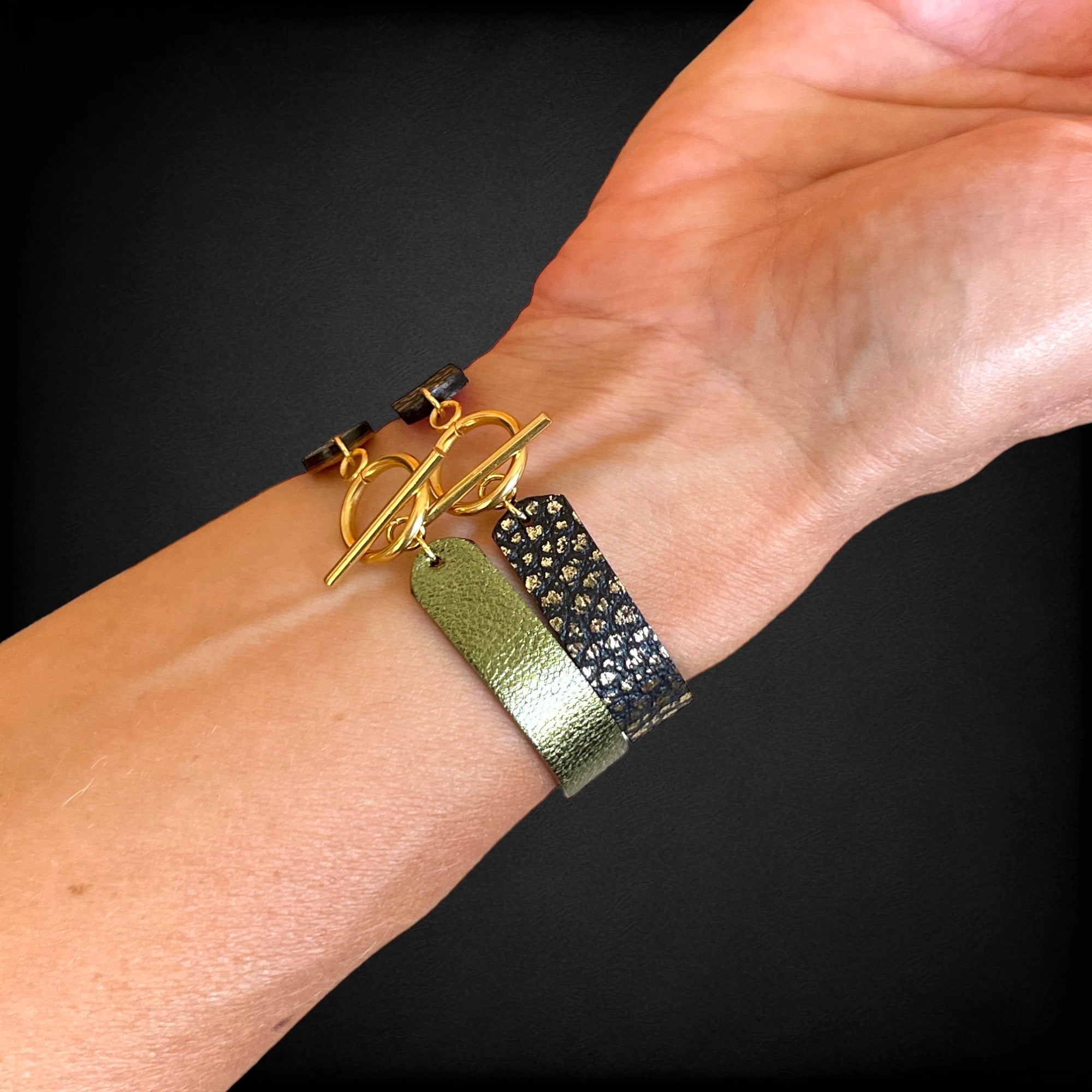 2 bracelets en cuir double face bicolores, avec fermoir boucle dorée en T, en cuirs noir grainé or pour le 1er, et kaki métallisé pour le second, portés au poignet et vus de dessous