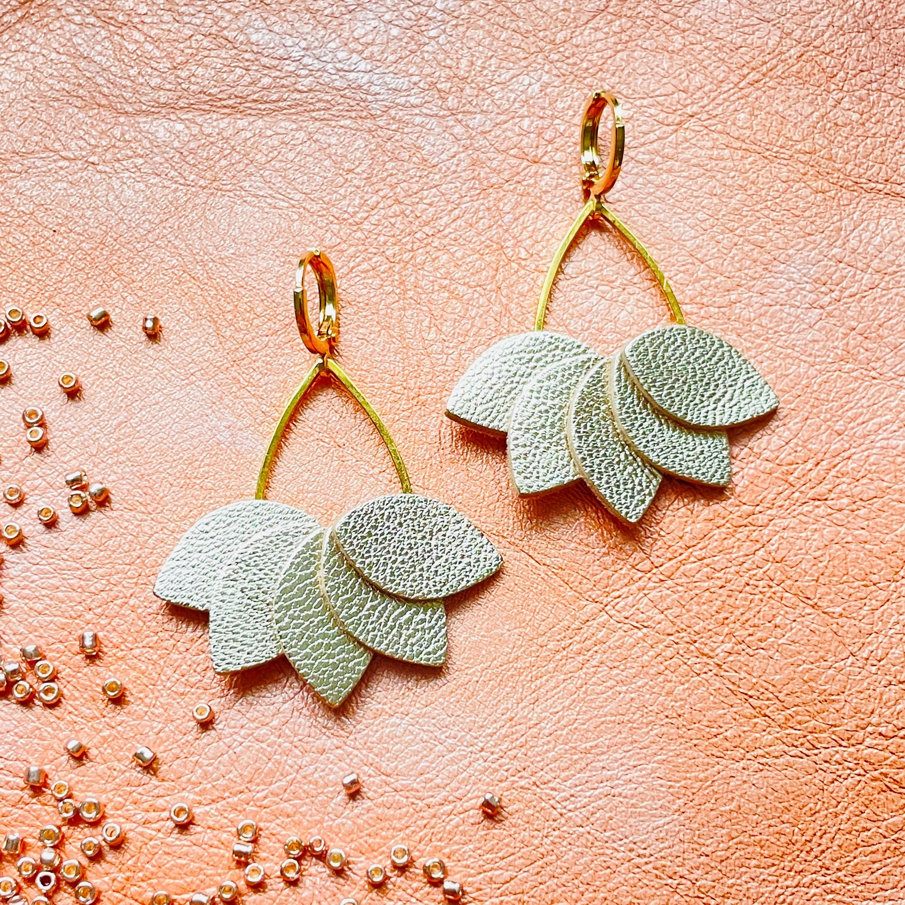 Boucles d'oreilles Nune modèle Aponi dorées à l'or fin avec pétales de cuir argent, en forme de couronne de pétales, vues de dessus avec perles de rocaille roses éparpillées, sur fond corail nacré clair