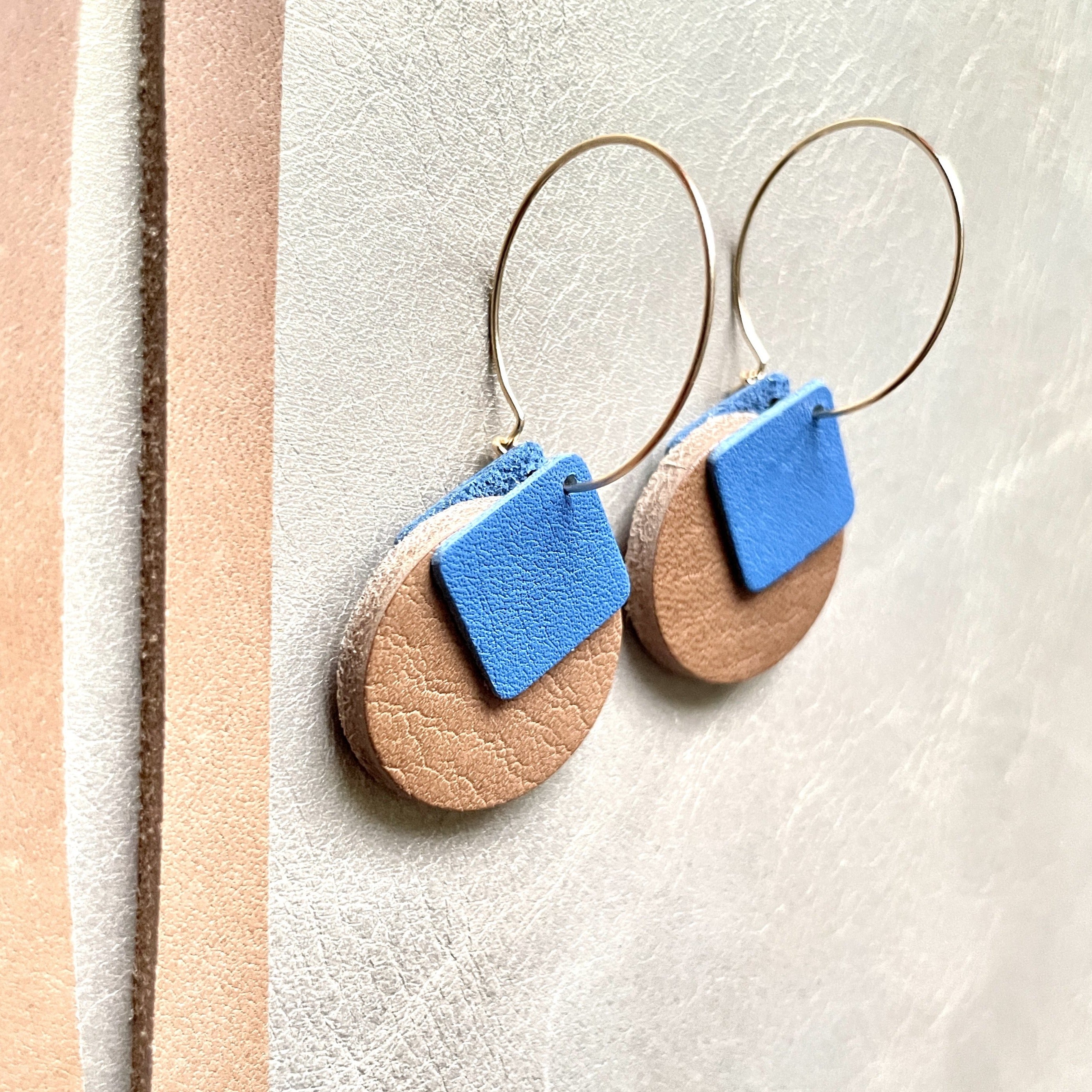boucles d'oreilles dorées Nune modèle Héra en cuirs beige amande et bleu denim mat sur fond gris doré, vues de côté
