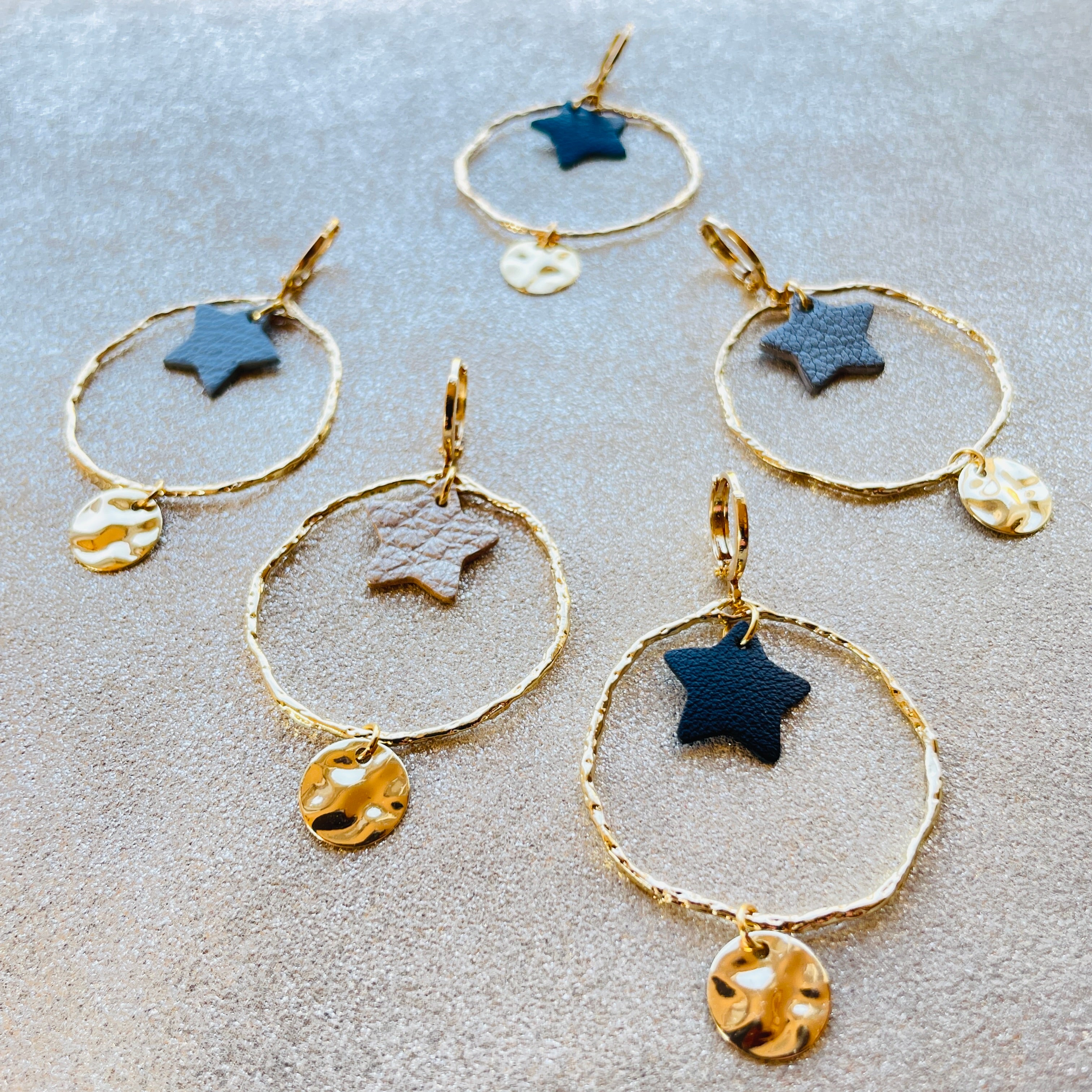 Boucles d'oreilles Nune modèle Naleli dorées à l'or fin de forme créole avec étoile de cuir coloré, vues de dessus, sur fond cuir beige pailleté