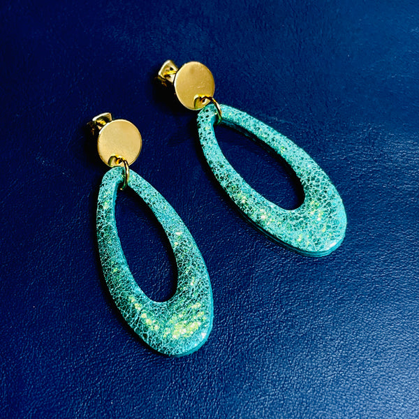 Puces d'oreilles dorées Drys de la marque Nune, avec leurs anneaux ovales en cuir vert émeraude brillant, sur fond cuir bleu marin satiné, vues de haut