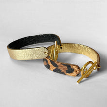 Charger l&#39;image dans la galerie, 2 bracelets réversibles à boucle fermoir en T en cuirs noir brillant façon serpent et doré antique mat et cuirs façon léopard et doré façon serpent, posés à plat l&#39;un sur l&#39;autre
