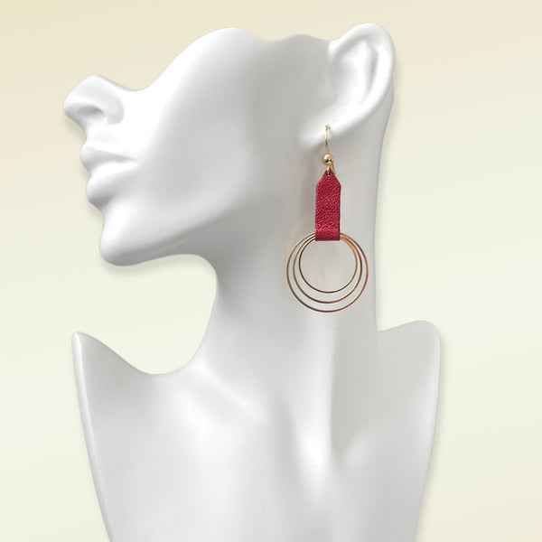 Grande boucle d'oreille dorée avec pendentif en anneaux concentriques attaché par un ruban de cuir recyclé rubis métallisé, portée à l'oreille. Modèle Dizzo