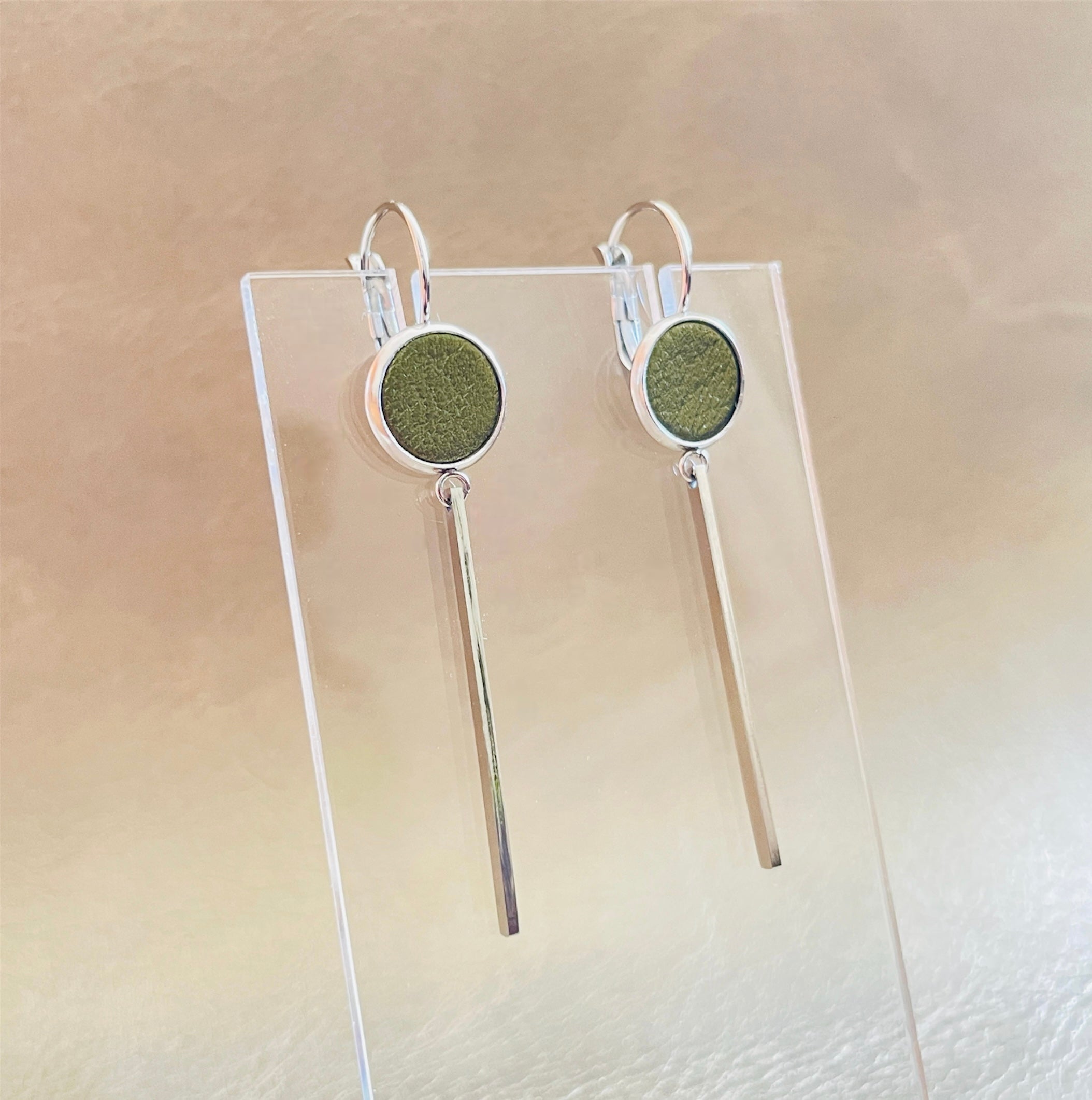 Longues et fines boucles d'oreilles argentées Naka avec cabochon en cuir kaki métallisé, sur fond beige