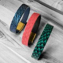 Charger l&#39;image dans la galerie, 3 bracelets en cuir marin rose saumon, bleu saphir métallisé et vert émeraude écaille avec fermoirs dorés aimantés, posés sur sur bois gris
