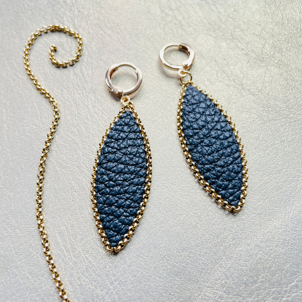 Paire de boucles d'oreilles pendantes Nune modèle Auris en forme d'épi en cuir noir grainé cerclé de chaîne dorée, sur fond cuir beige doré