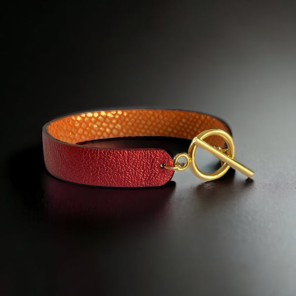 Bracelet en cuir double face bicolore, avec cuir rubis métallisé au recto et cuir orangé or façon croco au verso avec fermoir boucle dorée en T, sur fond noir