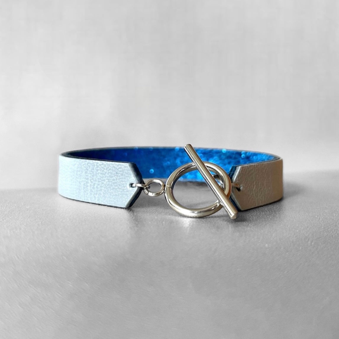 Bracelet en cuir double face bicolore, avec cuir bleu roi métallisé au verso et gris métallisé au recto avec fermoir boucle argentée en T, sur fond gris