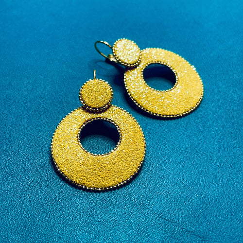 boucles d'oreilles créoles Nune modèle Céleste en forme de cercles en cuir safran pailleté argent, cerclées de chaîne dorée, sur fond cuir bleu canard