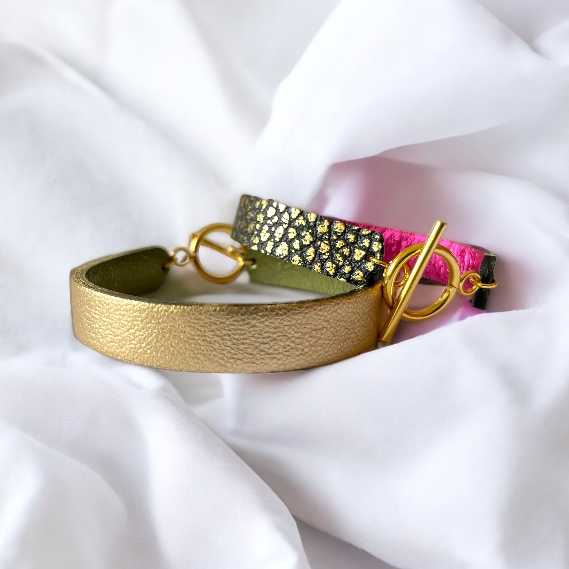 2 bracelets en cuir double face bicolores, avec fermoir boucle dorée en T, en cuirs fushia métallisé et noir grainé or pour le 1er, et doré antique mat et kaki métallisé pour le second, sur un linge blanc