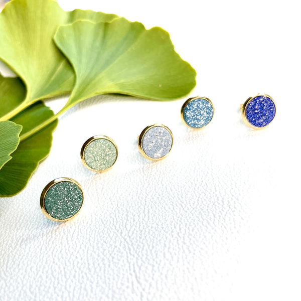 Puces d'oreilles dorées rondes Nune modèle Ilis dans 5 coloris de cuirs pailletés bleus et verts, sur fond blanc avec feuilles de Gingko