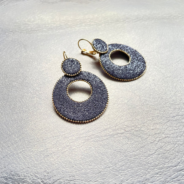 boucles d'oreilles créoles Nune modèle Céleste en forme de cercles en cuir anthracite pailleté argent, cerclées de chaîne dorée, sur fond beige doré