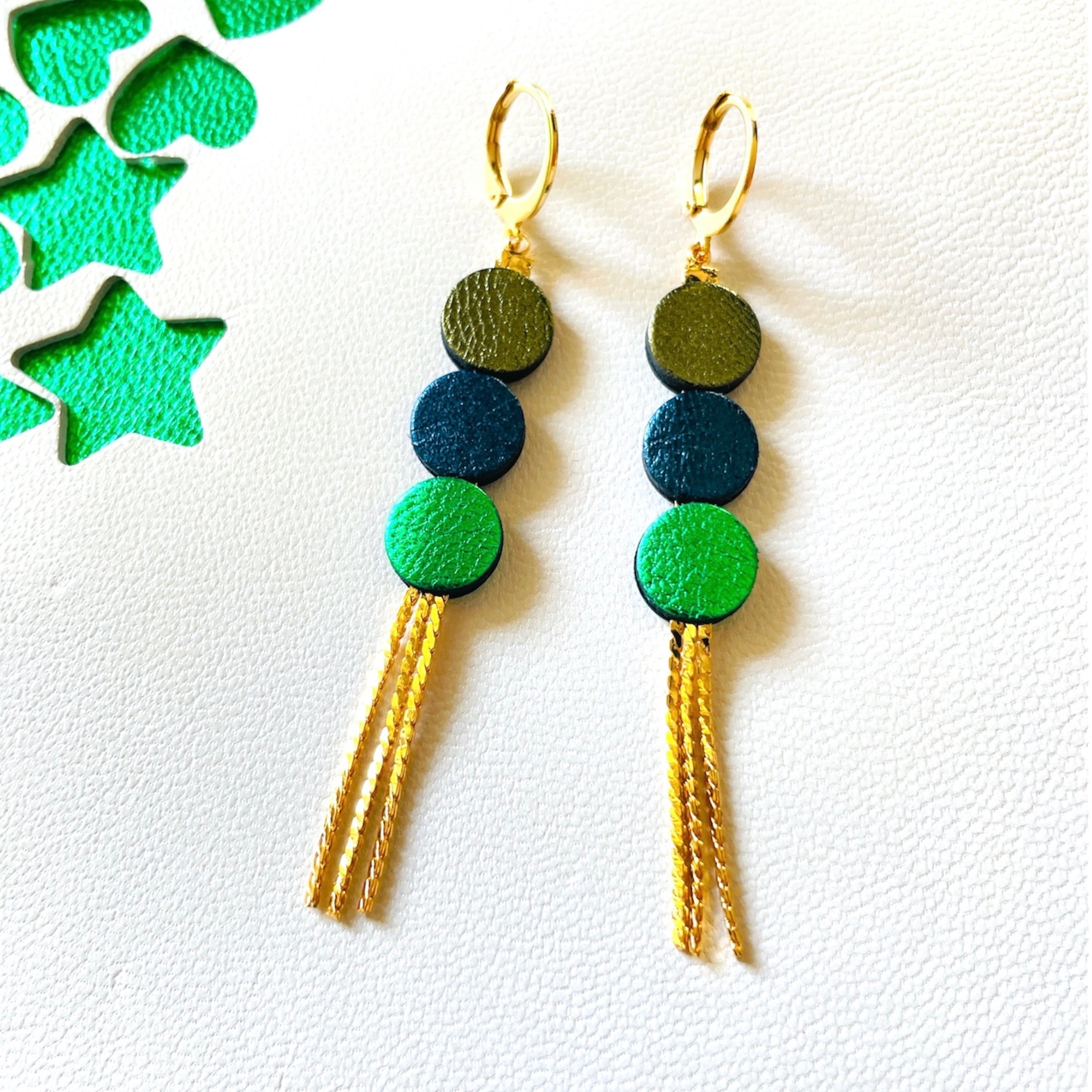 Longues et fines boucles d'oreilles Nune à franges dorées avec 3 petits disques de cuir recyclé kaki métallisé, bleu nuit métallisé et vert métallisé, sur fond blanc et vert étoilé. Modèle Janis