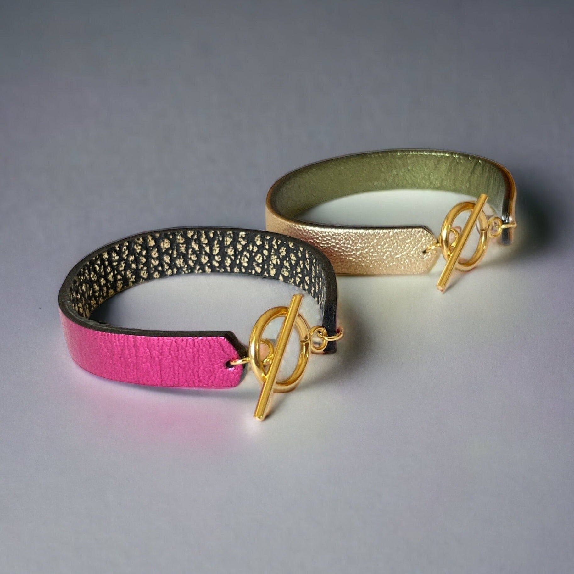 2 bracelets en cuir double face bicolores, avec fermoir boucle dorée en T, en cuirs fushia métallisé et noir grainé or pour le 1er, et doré antique mat et kaki métallisé pour le second, sur fond gris