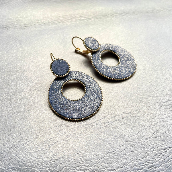 boucles d'oreilles créoles Nune modèle Céleste en forme de cercles en cuir gris pailleté or, cerclées de chaîne dorée, sur fond beige doré