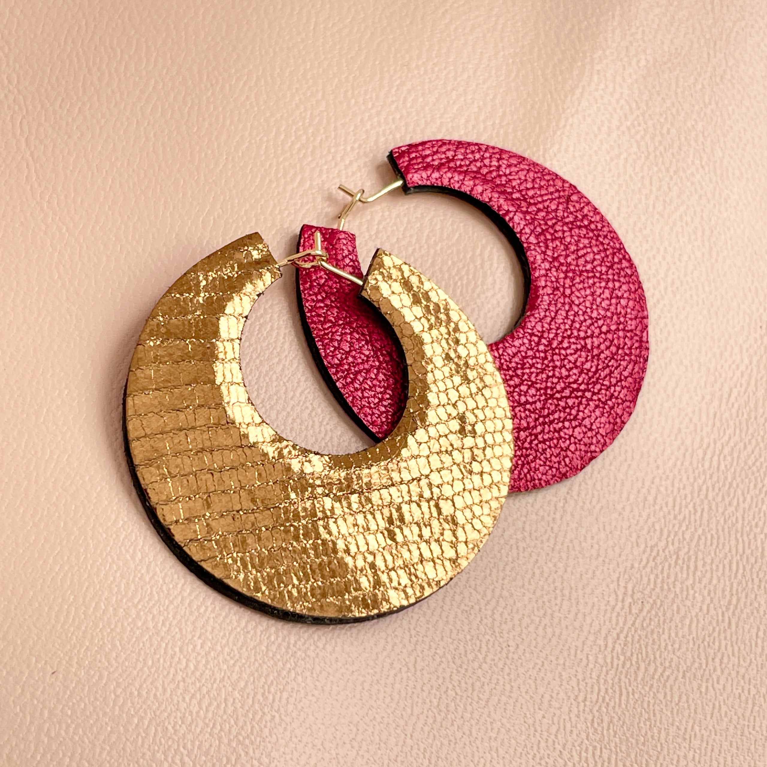 NUNE grandes boucles d'oreilles créoles dorées rondes tout-cuir Circus, un côté rubis métallisé, l'autre côté bronze brillant façon serpent, à plat sur fond beige rosé