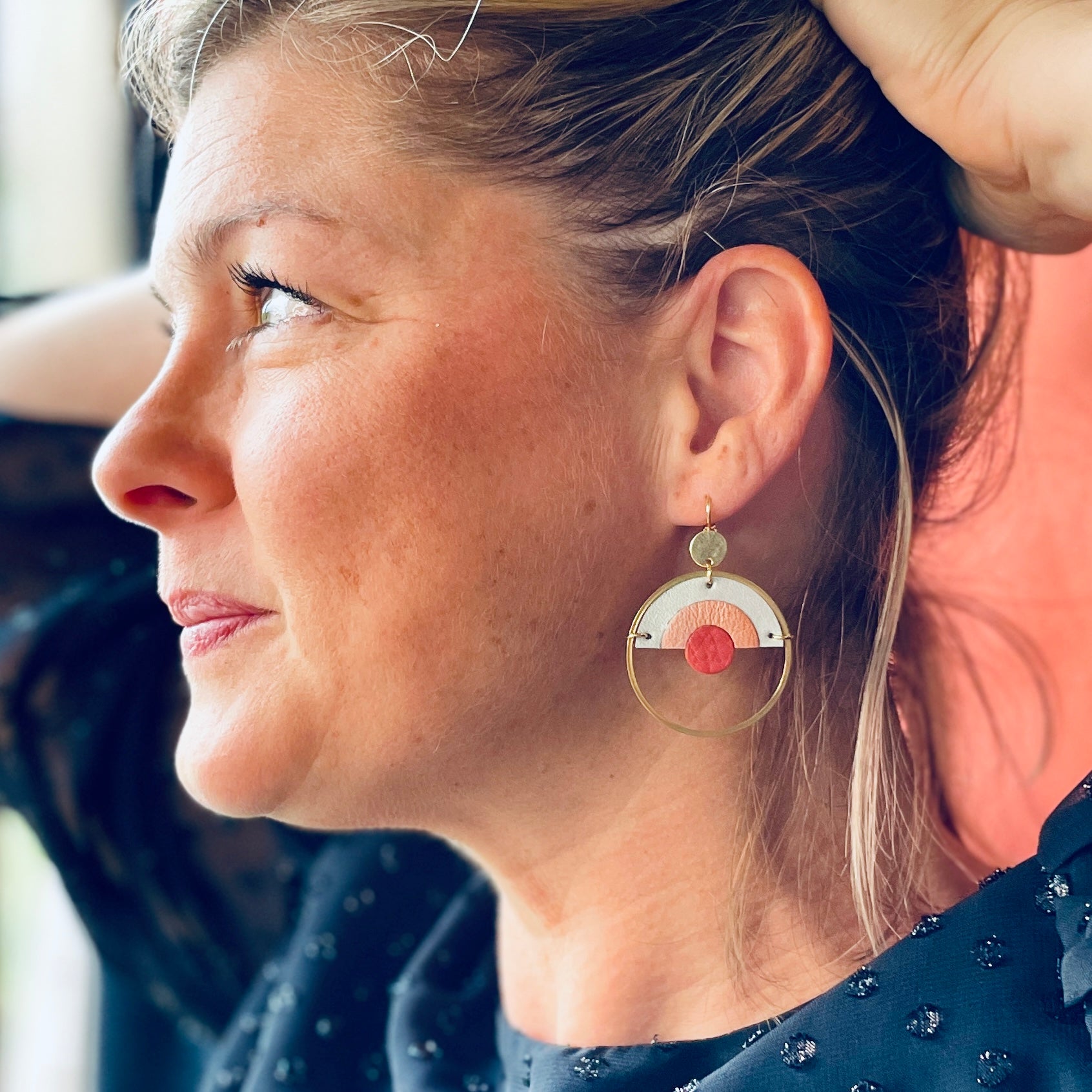 Boucle d'oreille dorée et ronde, modèle Sunset de la marque Nune, avec un dégradé arc-en-ciel de cuirs rose incarnat, corail nacré clair et bleu lune nacré, portée par un modèle aux cheveux blonds