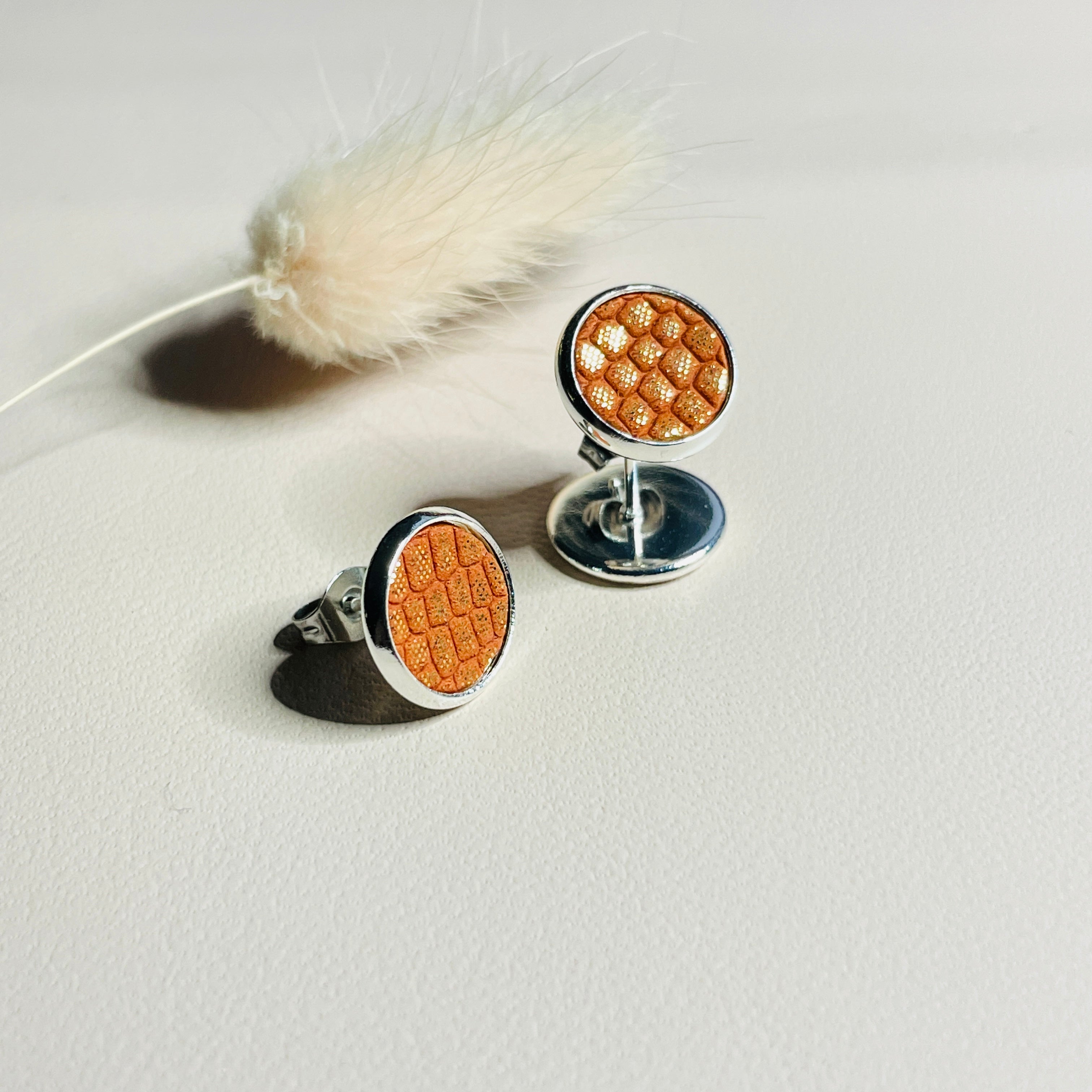 Puces d'oreilles argentées rondes Nune modèle Ilis argenté avec pastilles de cuir orangé or façon serpent, sur fond de cuir blanc cassé