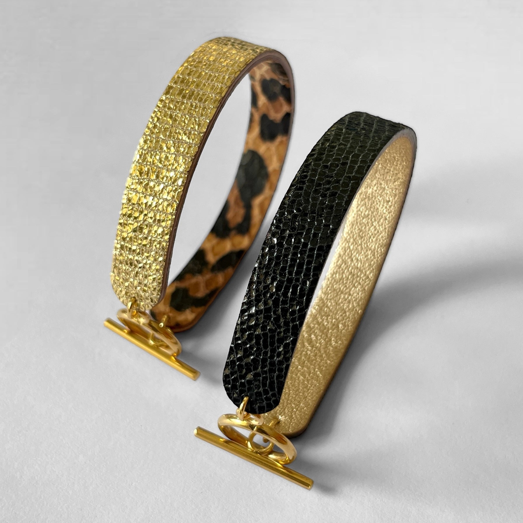 2 bracelets réversibles à boucle fermoir en T en cuirs noir brillant façon serpent et doré antique mat et cuirs façon léopard et doré façon serpent, posés sur la tranche