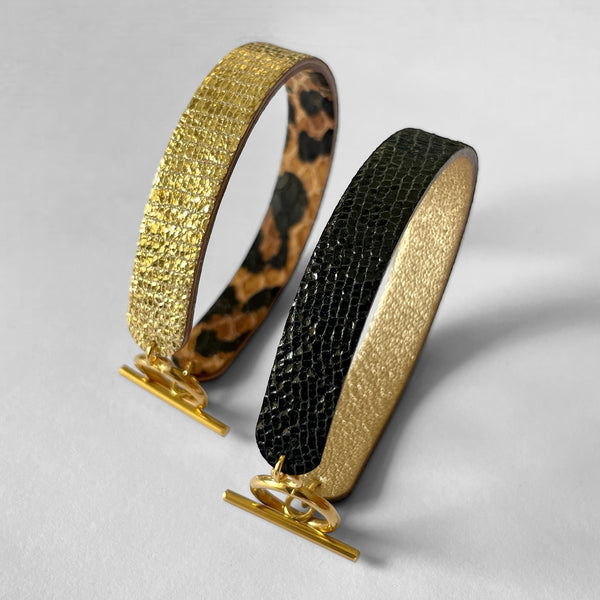 2 bracelets réversibles à boucle fermoir en T en cuirs noir brillant façon serpent et doré antique mat et cuirs façon léopard et doré façon serpent, posés sur la tranche