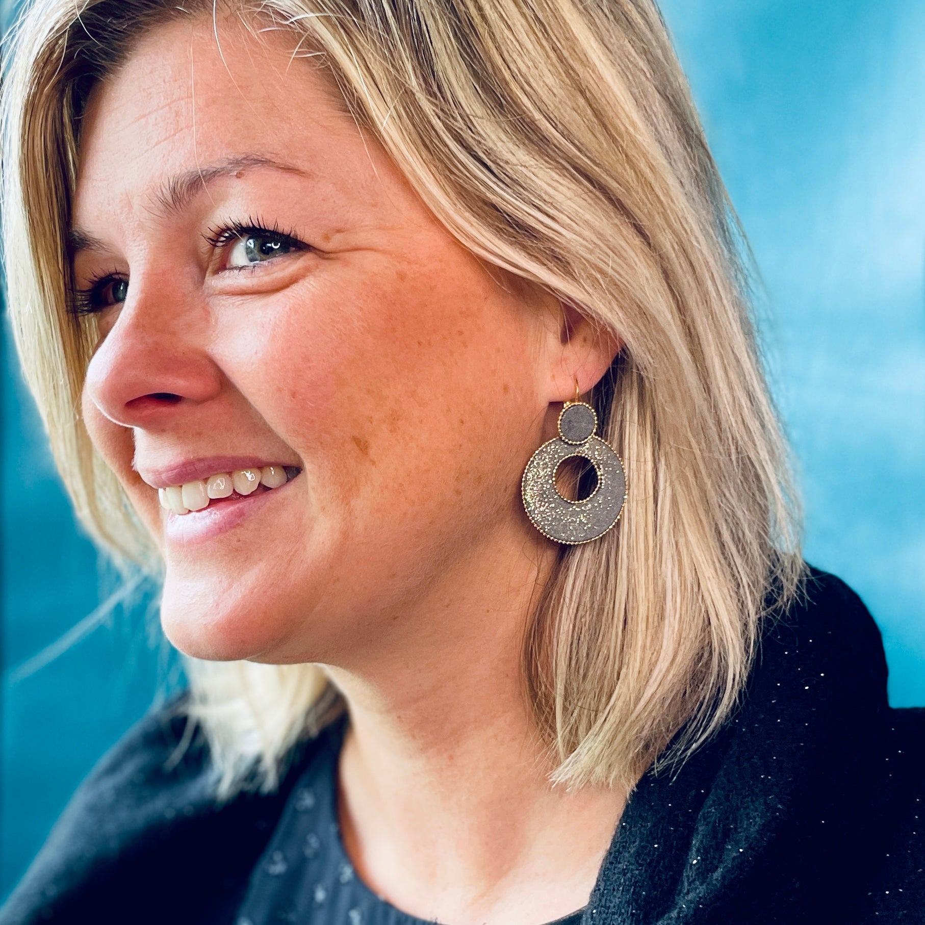 boucle d'oreille créole Nune modèle Céleste en forme de cercle en cuir gris pailleté or, cerclée de chaîne dorée, portée par un modèle aux cheveux blonds