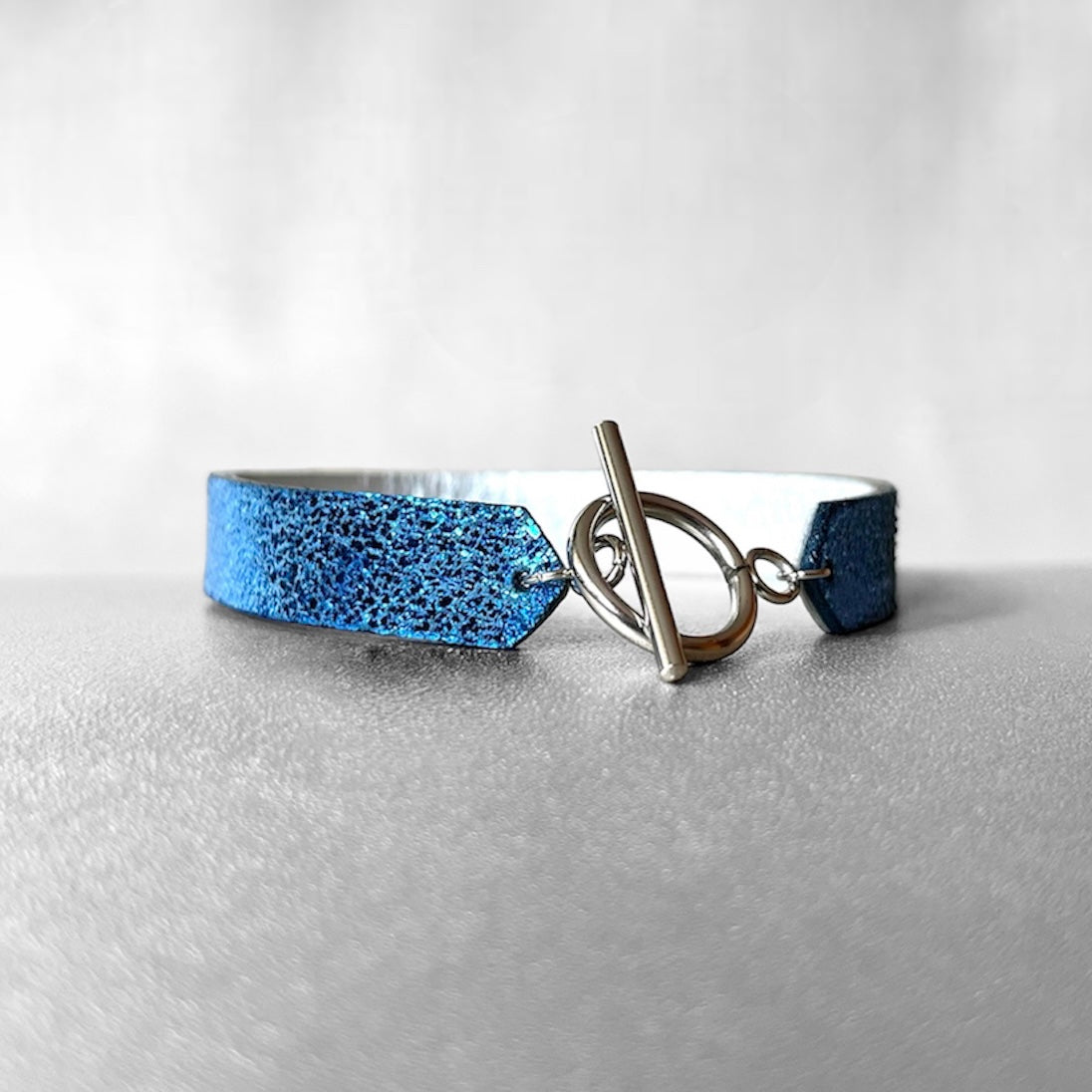 Bracelet en cuir double face bicolore, avec cuir bleu roi métallisé au recto et gris métallisé au verso avec fermoir boucle argentée en T, sur fond gris