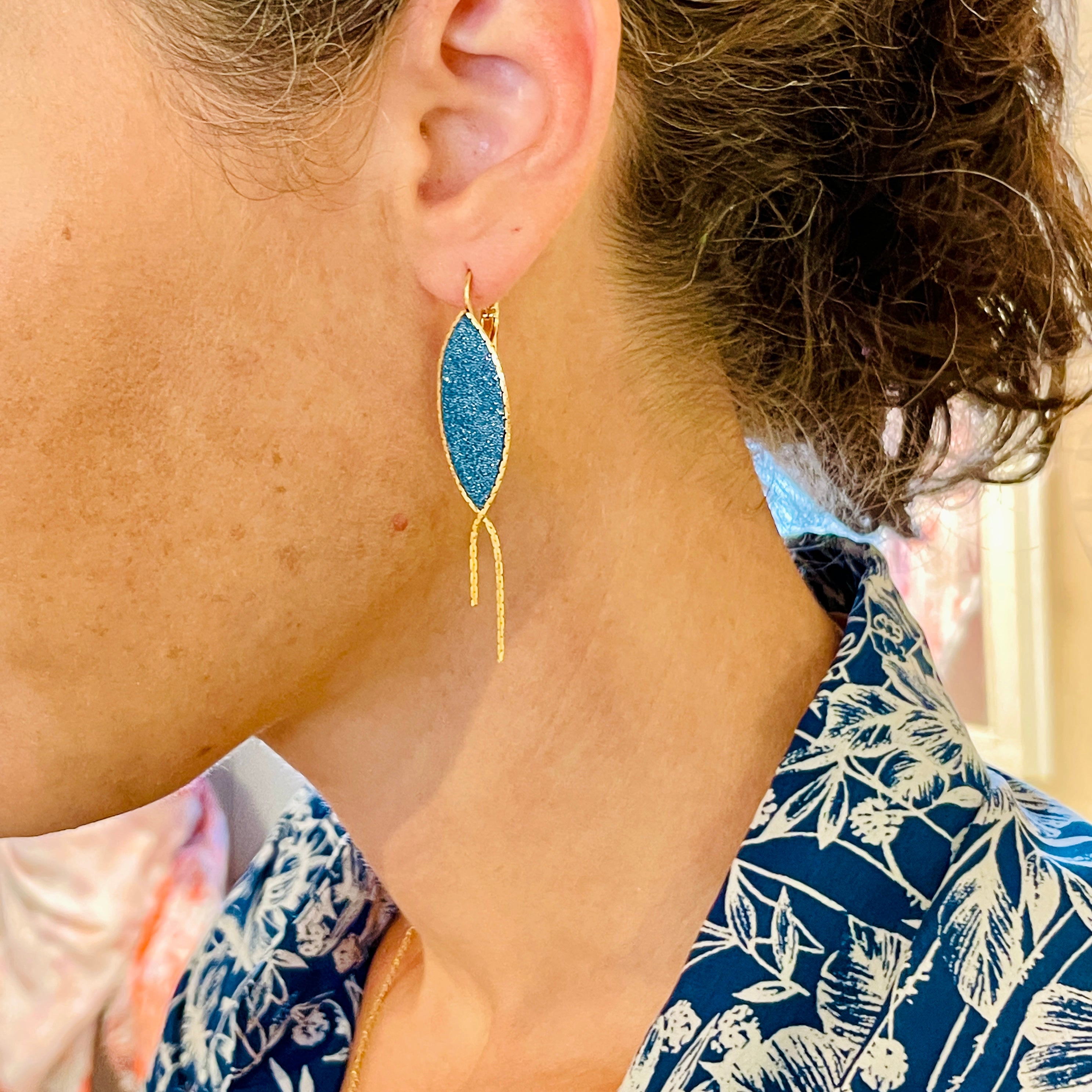 Boucle d'oreille Nune modèle Nayati en forme de fin poisson en cuir bleu paon pailleté argent, cerclé de chaîne dorée, portée à l'oreille