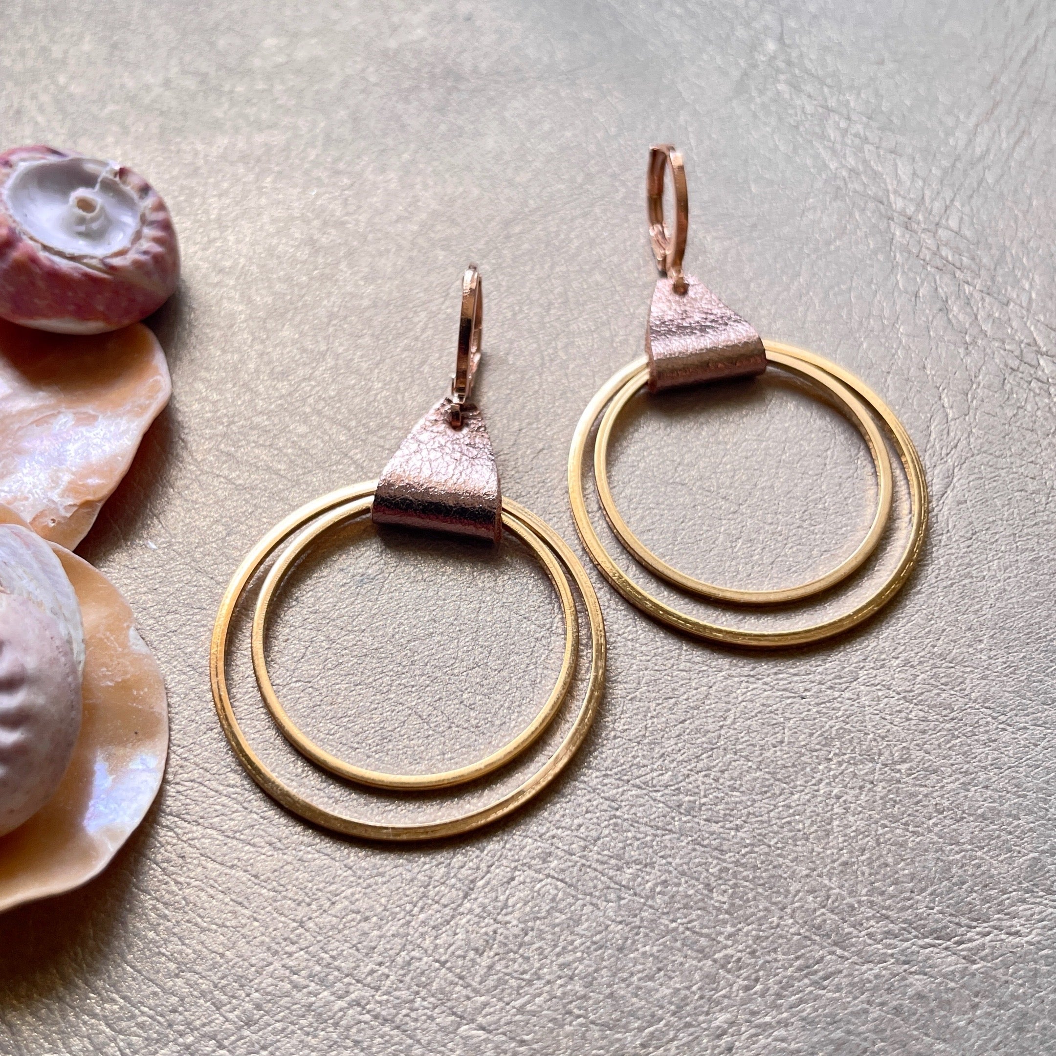 Créoles dorées du modèle Vega de la marque Nune, avec une touche de cuir cannelle dorée satinée, sur fond beige doré et coquillages 