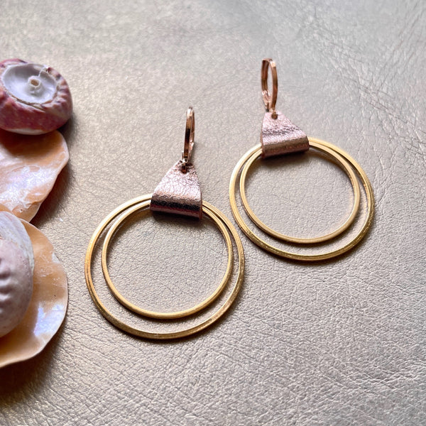 Créoles dorées du modèle Vega de la marque Nune, avec une touche de cuir cannelle dorée satinée, sur fond beige doré et coquillages 