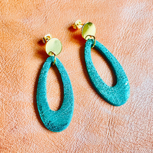 Puces d'oreilles dorées Drys de la marque Nune, avec leurs anneaux ovales en cuir vert émeraude brillant, sur fond cuir corail nacré clair, vues de haut