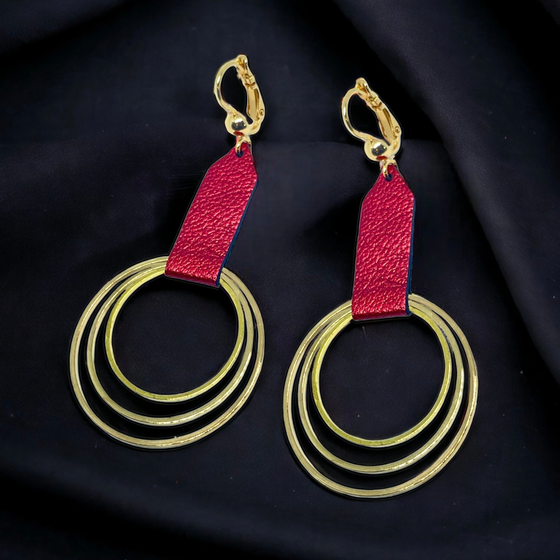 Grandes boucles d'oreilles dorées avec pendentifs en anneaux concentriques attachés par des rubans de cuir recyclé rubis métallisé, sur fond noir. Modèle Dizzo