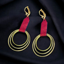 Charger l&#39;image dans la galerie, Grandes boucles d&#39;oreilles dorées avec pendentifs en anneaux concentriques attachés par des rubans de cuir recyclé rubis métallisé, sur fond noir. Modèle Dizzo
