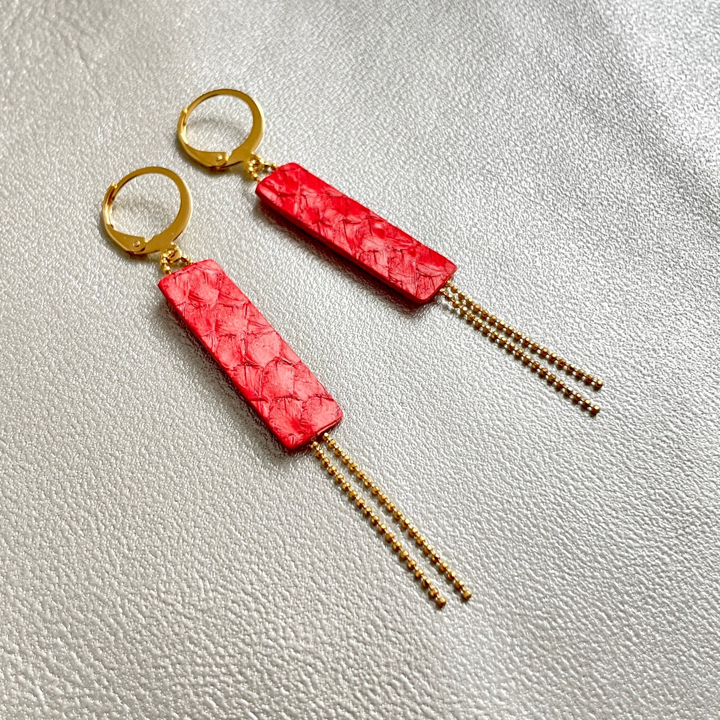 Boucles d'oreilles Effinis de Nune, dorées à l'or fin avec cuir marin, cuir de poisson saumon rouge cardinal, de forme effilée avec chaînettes dorées pendantes, sur fond cuir gris argenté