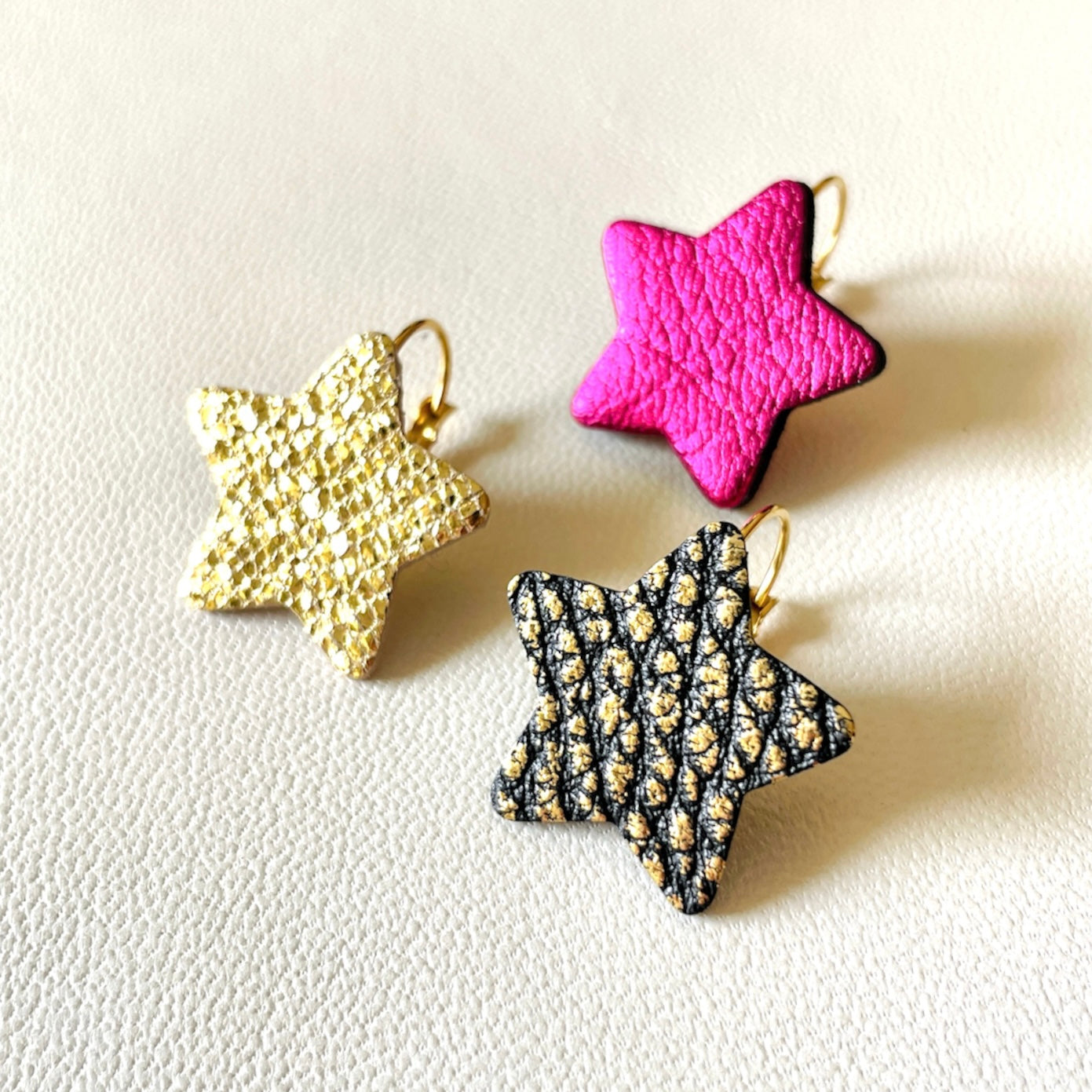 NUNE Boucles d'oreilles Diva en forme d'étoiles en cuir recyclé doré façon serpent, noir grainé or et fushia métallisé, sur fond blanc