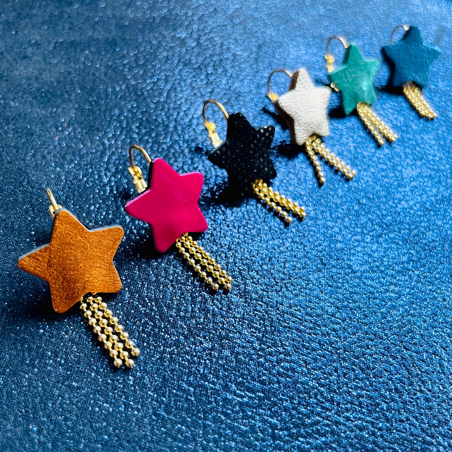 Rangée de boucles d'oreilles dorées Nune modèle Stella en forme d'étoiles, de différents coloris de cuirs recyclés : orange métallisé, rose fushia vernis, noir grainé irisé, argent, vert émeraude brillant, bleu roi métallisé